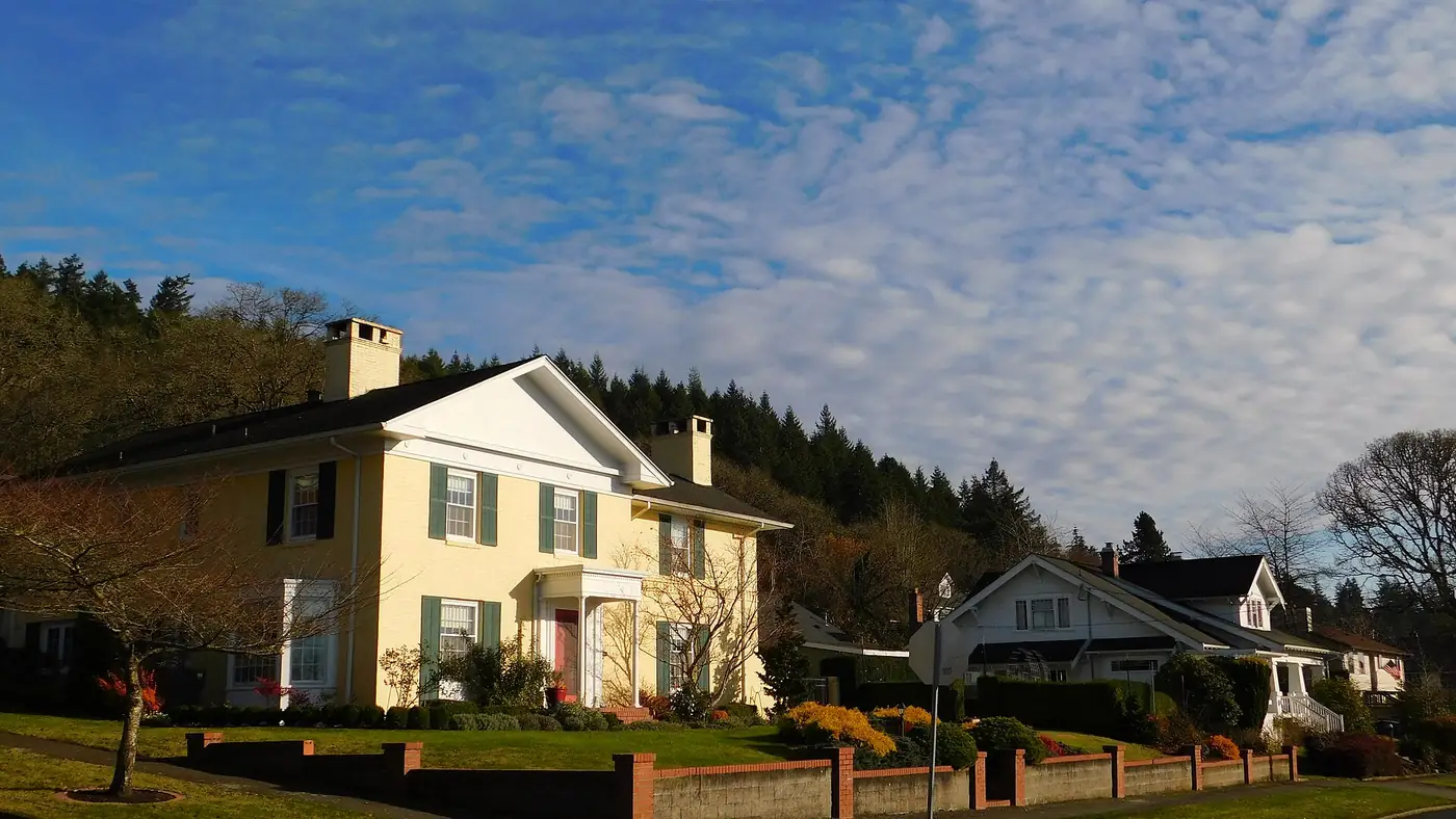 Pale yellow two-story house with a brick wall and a wooded hill behind it in Chehalis.