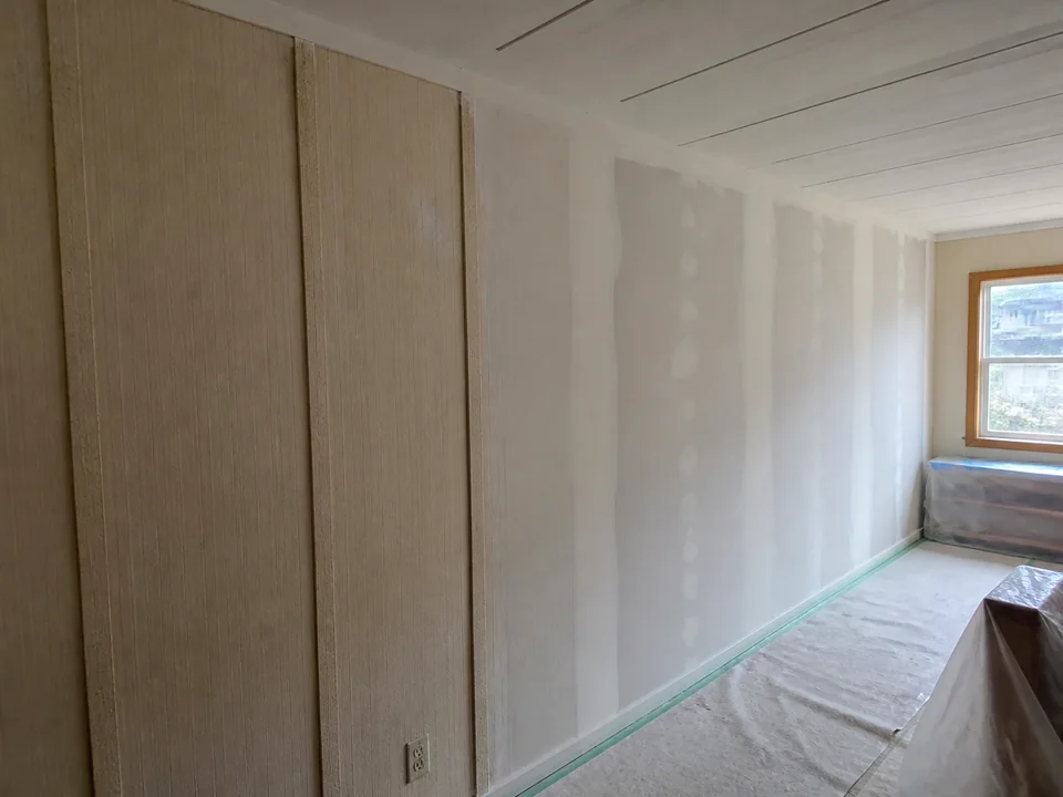 Manufactured-home wall transitioning from visible panel battens to a smooth skim-prepared surface